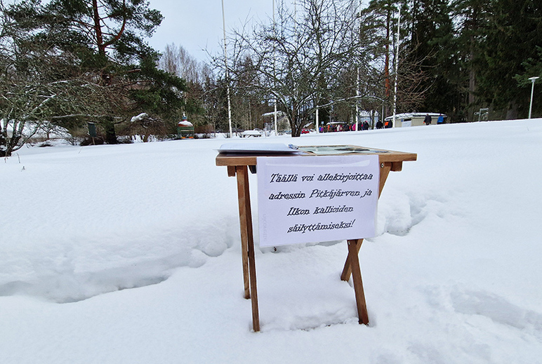 Lumisen kentän vieressä on pieni pöytä, jonka edessä paperissa lukee: Täällä voi allekirjoittaa adressin Pitkäjärven ja Ilkon kallioiden säilyttämiseksi!