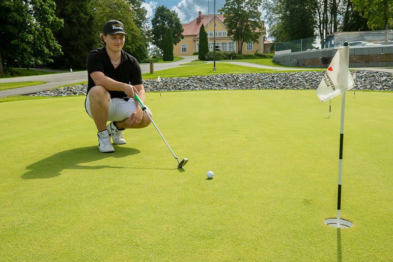 Lippalakkipäinen nuori mies on kyykyssä sileällä kentällä ja pitää käsissään golfmailaa. Hänen edessään on pieni pallo, ja kuvan reunassa lippu.