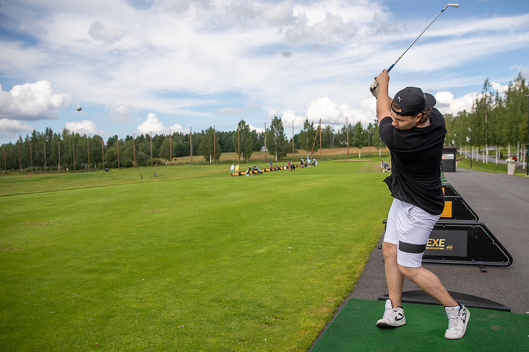 Lyöjän golfmaila on korkealla ilmassa, ja pallo lentää eteenpäin. Taustalla on laaja vihreä kenttä.