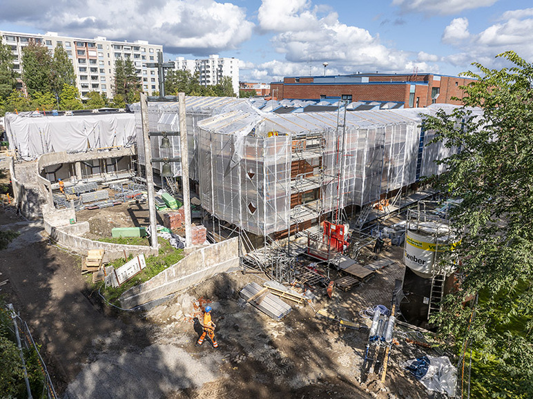 Iso rakennus on suojattu pressuilla, ja joka puolella on telineitä ja metalliputkia. Rakennuksen vierellä kävelee joku työhaalareissa.