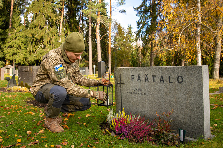 Maastotakkinen mies kyykyssä hautakiven vieressä syksyisellä hautausmaalla.