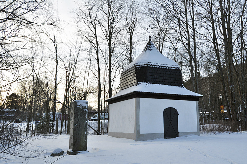 Pieni kappelirakennus, jossa on kellon muotoinen katto, ja sen vieressä kivipaasi, ovat lumisessa maisemassa. Taustalta kajastaa vähän auringonvaloa.