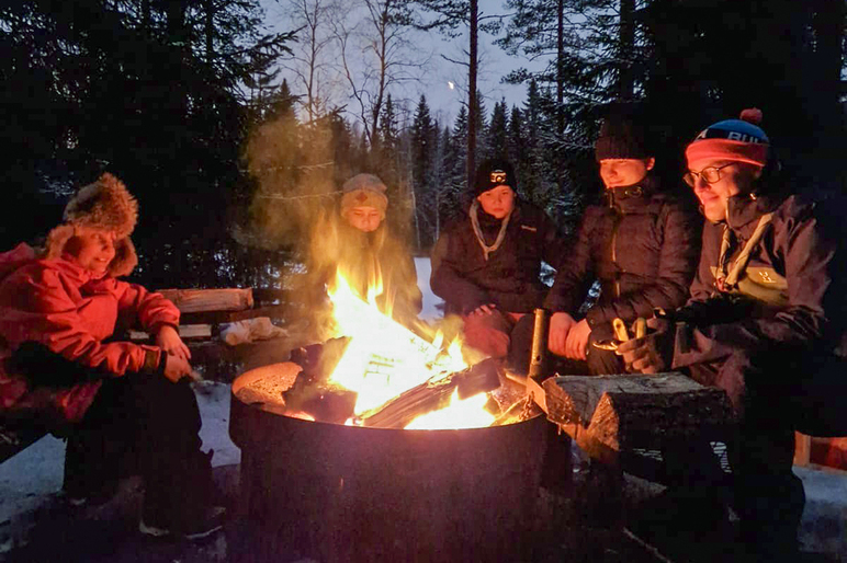 Partiolaiset istuvat leirinuotion äärellä hämärtyvässä illassa.